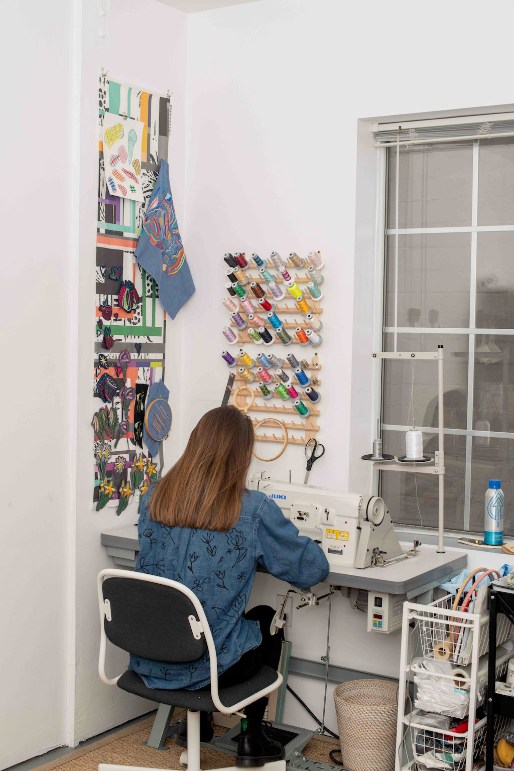 Photo of back of woman sat at her sewing machine machine in her making studio. Above her are different coloured threads on a wall spool holder.